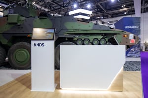 KNDS booth at DSEI 2025 featuring an armored vehicle in the foreground and a modern white reception counter.