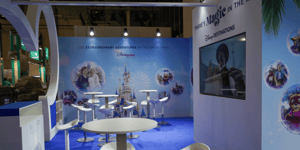 Detailansicht eines Bereichs des Disney-Messestands mit weißen Stehtischen und Hockern vor einer blau gestalteten Wand. Die Wand zeigt Disney-Motive in runden Grafikelementen und ein großes Schlossmotiv. Rechts ist ein Bildschirm in das Standdesign integriert.
