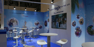 Innenansicht des Disney-Messestands mit mehreren weißen Stehtischen und Hockern. Die Rückwand zeigt das Motto ‚There’s Magic in the Air‘ sowie verschiedene Disney-Grafiken. Links befindet sich ein großer Monitor mit präsentierten Inhalten.