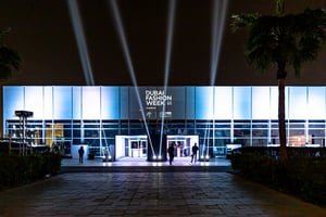 Nighttime exterior of a modern building illuminated with bright lights, featuring a large ‘Dubai Fashion Week’ sign and sponsor logos.