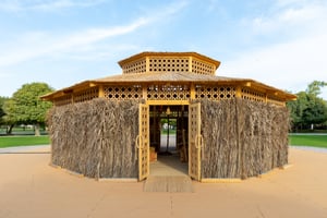 A traditional hut made of wood and straw in an open area with trees and grass in the background.