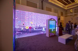 Exhibition featuring a large digital installation displaying colorful pixel art, viewed by several people.