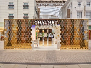 Der Pop-up-Laden von CHAUMET besticht durch sein auffälliges Design mit gold- und silberfarbenen, verspiegelten Sechseckpaneelen. Das Logo befindet sich oben in der Mitte, im Inneren stehen elegante Sitzgelegenheiten und Tische bereit.