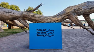 Large piece of driftwood or tree trunk on the ground, with a blue sign in front reading “Public Art Abu Dhabi Biennial” in English and Arabic.