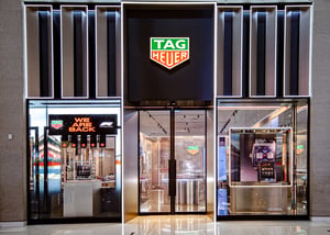 TAG Heuer storefront with illuminated logo, glass facade, and display windows showcasing watches and promotional materials. A sign reading ‘WE ARE BACK’ with an F1 logo is visible.