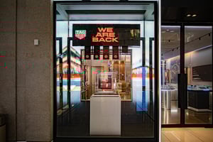 TAG Heuer Ladenfront mit beleuchtetem Logo und großem Schild ‚WE ARE BACK‘ über dem Eingang. Innen sind Uhren und Displays sichtbar.