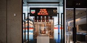 TAG Heuer Ladenfront mit beleuchtetem Logo und großem Schild ‚WE ARE BACK‘ über dem Eingang. Innen sind Uhren und Displays sichtbar.