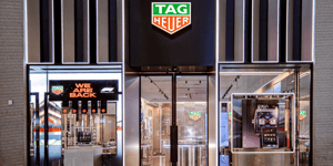 TAG Heuer-Ladenfront mit beleuchtetem Logo, Glasfassade und Schaufenstern, in denen Uhren und Werbematerialien ausgestellt sind. Ein Schild mit der Aufschrift „WE ARE BACK” und einem F1-Logo ist zu sehen.