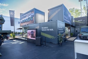 Außenansicht des Mercedes-Benz Defence Trucks Messestands aus übereinandergestapelten Containermodulen mit großflächigen Fahrzeuggrafiken und digitalen Präsentationsflächen.