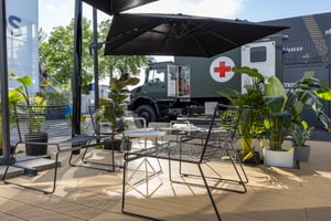 Terrassenbereich des Messestands mit Sitzgelegenheiten, Pflanzen und Blick auf ein ausgestelltes Militärfahrzeug von Mercedes-Benz Defence Trucks.