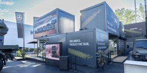 Außenansicht des Mercedes-Benz Defence Trucks Messestands aus übereinandergestapelten Containermodulen mit großflächigen Fahrzeuggrafiken und digitalen Präsentationsflächen.