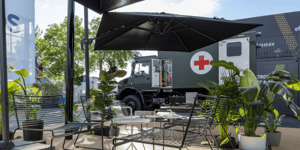 Terrassenbereich des Messestands mit Sitzgelegenheiten, Pflanzen und Blick auf ein ausgestelltes Militärfahrzeug von Mercedes-Benz Defence Trucks.