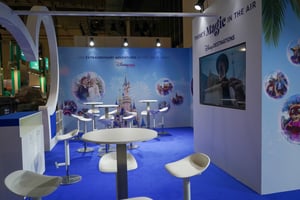 Close-up view of a Disney exhibition booth section with white high tables and stools in front of a blue wall. The wall features circular Disney-themed illustrations and a prominent castle graphic. A screen is integrated into the right side of the booth.