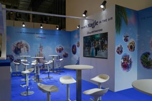 Interior view of the Disney booth featuring several white high tables and stools. The back wall displays the slogan ‘There’s Magic in the Air’ along with multiple Disney-themed graphics. A large screen on the left side shows visual content.
