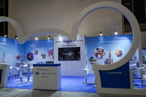 View of the Disney exhibition booth featuring a large white Mickey Mouse outline in the foreground. The booth includes a presentation screen, several high tables, and blue walls decorated with Disney graphics and a central castle illustration.