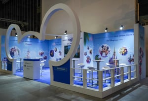 Open Disney exhibition booth featuring a blue floor, white wall structures, and a large curved Mickey Mouse silhouette. Several high tables with white stools are arranged in front of a backdrop decorated with Disney-themed graphics and a central castle illustration.