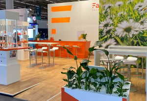 A view of the Faller Packaging stand at CPHI 2025, featuring a dandelion pattern on the walls, glass display areas and orange design elements.
