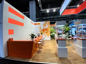 Faller Packaging exhibition stand at CPHI Frankfurt with orange design elements, a counter with bar stools and glass displays.
