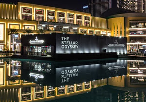 Ein beleuchtetes Gebäude bei Nacht mit der Aufschrift „THE STELLAR ODYSSEY“ und „Jaeger-LeCoultre“. Die Spiegelung des Gebäudes ist im davorliegenden Wasser sichtbar und erzeugt einen symmetrischen Effekt.
