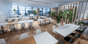 Heller Hospitality-Bereich im Innern des KNDS-Gebäudes mit weißen Tischen, hellen Stühlen, Holzelementen und Grünpflanzen, die eine offene und einladende Atmosphäre schaffen.