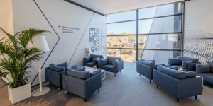 Ruhiger Lounge-Bereich im Obergeschoss mit blauen Sesseln, einer Grünpflanze und großflächiger Verglasung; an der Wand der Schriftzug „FROM A HIGHER GROUND: DISCOVER OUR LAND DEFENSE