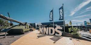 Großer dreidimensionaler KNDS-Schriftzug auf einer Holzdeckfläche im Außenbereich der Eurosatory 2026, flankiert von KNDS-Flaggen und militärischen Großexponaten.