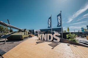 Großer dreidimensionaler KNDS-Schriftzug auf einer Holzdeckfläche im Außenbereich der Eurosatory 2026, flankiert von KNDS-Flaggen und militärischen Großexponaten.