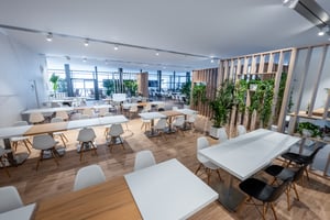Heller Hospitality-Bereich im Innern des KNDS-Gebäudes mit weißen Tischen, hellen Stühlen, Holzelementen und Grünpflanzen, die eine offene und einladende Atmosphäre schaffen.