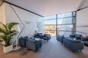 Ruhiger Lounge-Bereich im Obergeschoss mit blauen Sesseln, einer Grünpflanze und großflächiger Verglasung; an der Wand der Schriftzug „FROM A HIGHER GROUND: DISCOVER OUR LAND DEFENSE
