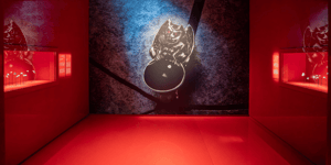An exhibition room with red lighting displays a large jewelry piece or artifact against a textured backdrop.