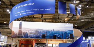 Main view of the Lufthansa Group stand at the ITB tourism trade fair, featuring a large ring-shaped structure on the ceiling and a large LED display.