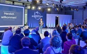 Ein Redner steht auf einer Bühne und begrüßt das Publikum. Im Hintergrund ist ein großer Bildschirm mit der Aufschrift „Herzlich willkommen!“ zu sehen.
