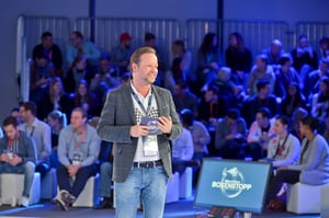 Ein Redner steht mit Mikrofon vor dem Publikum und spricht. Die Zuhörer sitzen und hören aufmerksam zu.