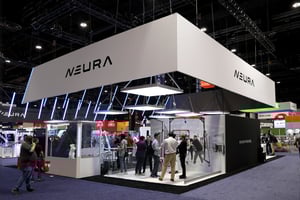 Innenansicht des NEURA Robotics Messestandes mit Glaswänden und Robotik-Demonstrationen. Mehrere Personen beobachten die Präsentationen.