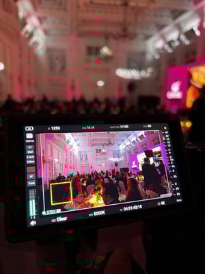 Blick durch einen Kamerabildschirm auf den Veranstaltungsraum, der mit Gästen gefüllt ist. Im Fokus steht die Bühne und deren Aufbau.