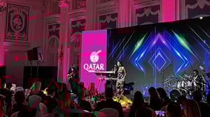Eine Bühne mit musizierenden Künstlern, davor ein Publikum an festlich gedeckten Tischen mit Kerzen. Im Hintergrund ist ein großer Bildschirm mit geometrischen Mustern und dem Logo von Qatar Airways zu sehen.