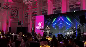 A stage with musicians performing, with an audience seated at candlelit tables. In the background, a large screen displays geometric patterns and the Qatar Airways logo.