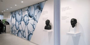 Ein Ausstellungsraum mit zwei Büsten und einer Wand mit einem wissenschaftlichen Muster. Die Büsten sind mit „OTTO SCHOTT“ und „ERNST ABBE“ beschriftet.