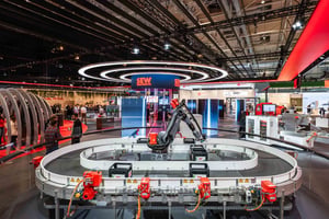 Automatisierte Industrieanlage mit Roboterarm im Mittelpunkt eines runden Förderbandsystems unter großen Ringlichtern am SEW‑Messestand.