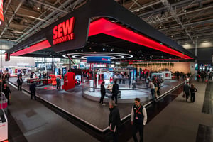 Side view of SEW-EURODRIVE's exhibition booth at Hannover Messe 2026 with a black ceiling, red highlights and logo, several visitors and a prominent circular design element in the center of the stand.