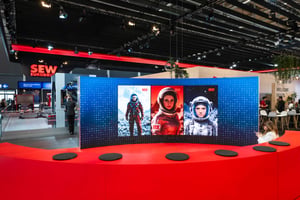 Gebogene LED‑Videowand mit drei Darstellungen von Raumanzug-Figuren auf roter Bühne am SEW‑Messestand, umgeben von Ausstellungsflächen.