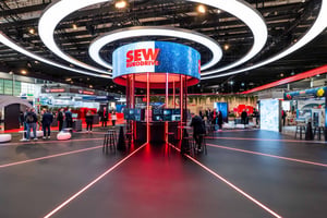 Moderner SEW‑EURODRIVE Messestand mit zentralem runden LED-Ring, beleuchtetem Kernpavillon und markantem Rot‑Schwarz-Design in einer großen Messehalle.