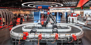 Automatisierte Industrieanlage mit Roboterarm im Mittelpunkt eines runden Förderbandsystems unter großen Ringlichtern am SEW‑Messestand.