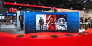 Gebogene LED‑Videowand mit drei Darstellungen von Raumanzug-Figuren auf roter Bühne am SEW‑Messestand, umgeben von Ausstellungsflächen.
