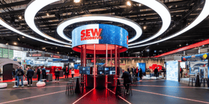 Moderner SEW‑EURODRIVE Messestand mit zentralem runden LED-Ring, beleuchtetem Kernpavillon und markantem Rot‑Schwarz-Design in einer großen Messehalle.