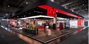 SEW-EURODRIVE booth at HANNOVER MESSE 2024 featuring a large red frame and circular exhibit in the foreground. Visitors are exploring the display area with interactive elements.