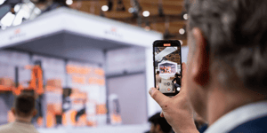 Nahaufnahme eines Besuchers, der den STILL-Messestand fotografiert. Im Hintergrund ist ein großes Display mit Regalsystemen und Lagertechnik zu sehen.