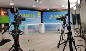 Ein Fernsehstudio mit mehreren Kameras und Beleuchtungseinrichtungen. Im Hintergrund ist eine blaue Wand mit der Aufschrift „JA SAGEN UND CO2 AUSGLEICHEN“ zu sehen. Zwei weiße Tische stehen vor der Wand.