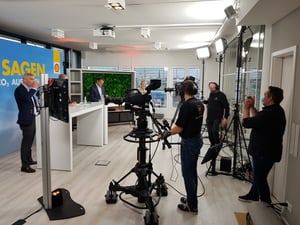 Das gleiche Fernsehstudio, aber diesmal sind mehrere Personen im Bild zu sehen, darunter Kameraleute und Moderatoren. Die blaue Wand mit der Aufschrift „JA SAGEN UND CO2 AUSGLEICHEN“ ist ebenfalls sichtbar.
