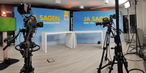 A television studio with several cameras and lighting equipment. In the background, there is a blue wall with the text 